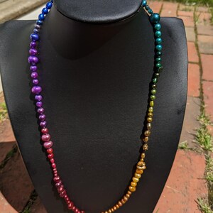 Pode incluir: Um colar de p&eacute;rolas iridescentes arco-&iacute;ris com um degrad&ecirc; de cores que vai do azul ao roxo, ao vermelho e ao dourado. O colar &eacute; exibido em um manequim preto.