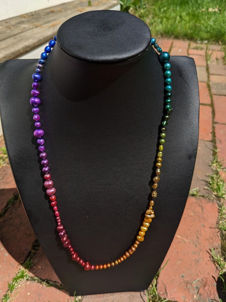 Pode incluir: Um colar de p&eacute;rolas iridescentes arco-&iacute;ris com um degrad&ecirc; de cores que vai do azul ao roxo, ao vermelho e ao dourado. O colar &eacute; exibido em um manequim preto.