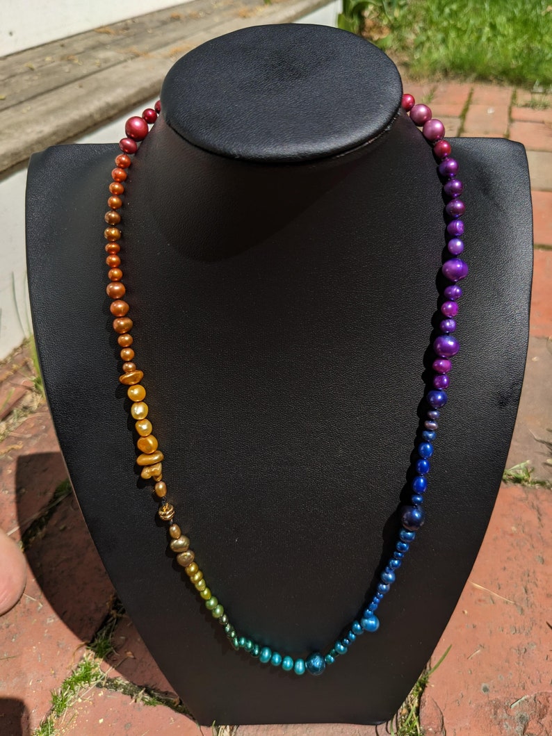 Pode incluir: Um colar degrad&ecirc; arco-&iacute;ris feito de contas iridescentes. O colar est&aacute; em um busto de manequim preto.