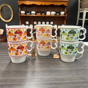 Puede incluir: Tres pilas de tazas de cerámica con diseños de paraguas y flores. Las tazas tienen bases blancas y asas en forma de corazón. Los diseños de paraguas presentan colores naranja, rosa y verde. Las tazas están apiladas sobre una superficie de madera oscura.