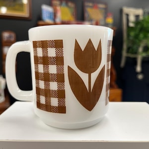 Puede incluir: Taza de cerámica blanca con un diseño de tulipán marrón y un patrón de cuadros marrones. La taza tiene un asa grande. La taza es de estilo vintage y está sobre un fondo borroso.