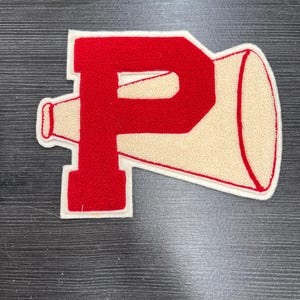 May include: A red and cream-colored felt patch in the shape of the letter "P" combined with a megaphone. The letter "P" is in red, with a cream-colored megaphone shape extending to the right. The patch has a white border.
