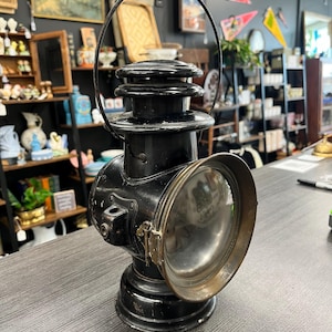 May include: A black metal railroad lantern featuring a circular glass lens with a brass-toned rim, a tiered top section, and a curved wire handle. The lantern has a sturdy base and a side-mounted attachment point, showing signs of wear.