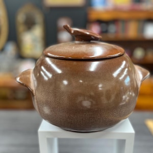 May include: A brown ceramic casserole dish with a lid. The dish has a rounded shape with two handles and a small decorative element on the lid. The dish is sitting on a white stand.