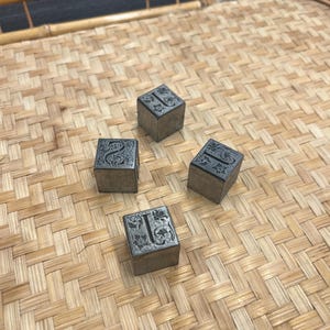 May include: Four metallic square blocks featuring embossed letters and intricate floral patterns on their top surfaces. Each block displays a distinct character, including the letters I, S, and L, with a dark, weathered finish on the sides and top.
