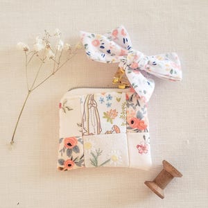 May include: A small square fabric pouch featuring a patchwork design of floral patterns and a religious figure illustration. A matching fabric bow is attached to the gold zipper pull. A sprig of dried white flowers and a wooden spool are nearby.
