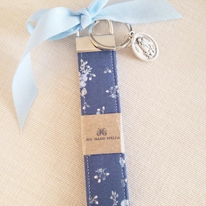 May include: A blue floral keychain wristlet with a silver key ring and a religious medal. The wristlet has a blue ribbon tied at the top. The text "AVE MARIS STELLA" is visible on the wristlet.