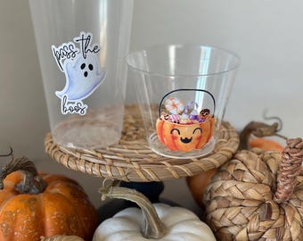 May include: Clear plastic cups with Halloween-themed stickers. One cup has a ghost with the text "pass the boos", and the other has a smiling jack-o'-lantern basket filled with candy. Pumpkins and gourds are arranged around the cups.