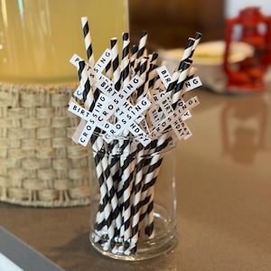 Könnte beinhalten: Ein klares Glasgefäß gefüllt mit schwarz-weiß gestreiften Papierstrohhalmen. Kleine weiße Etiketten mit den Wörtern "BIRTHDAY", "CROSSING" und "SPORTS" sind an den Strohhalmen befestigt. Im Hintergrund befindet sich ein großer Getränkespender aus Glas.
