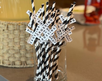 May include: A clear glass jar filled with black and white striped paper straws. Small white tags with the words "BIRTHDAY", "CROSSING", and "SPORTS" are attached to the straws. A large glass beverage dispenser is in the background.