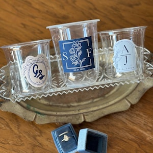 Op de afbeelding: Drie doorzichtige plastic bekers met gepersonaliseerde stickers, gerangschikt op een decoratief dienblad. De stickers bevatten initialen, bloemmotieven en data. Twee blauwe fluwelen ringendoosjes zijn ook aanwezig, waarvan één open met een diamanten ring.