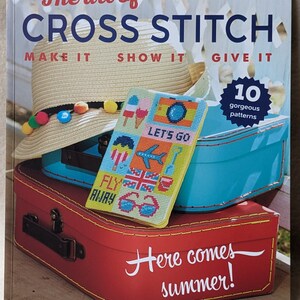Może przedstawiać: Okładka książki z tytułem "The art of CROSS STITCH MAKE IT SHOW IT GIVE IT" i kolorową ilustracją walizki, kapelusza i projektu haftu krzyżykowego w letnim stylu. Na okładce znajduje się również tekst "Here comes summer!"