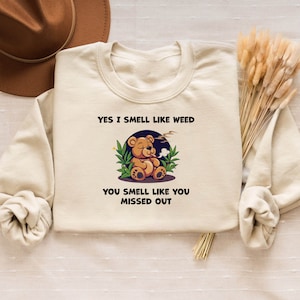 May include: A beige sweatshirt with the text "Yes I smell like weed" and a cartoon bear smoking a joint. The text below the bear says "You smell like you missed out."