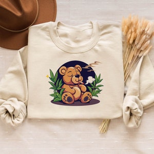Peut inclure: Un sweat-shirt beige avec une illustration de dessin animé d'un ours en peluche brun portant un bandana vert et fumant un joint. L'ours est entouré de feuilles de marijuana vertes.