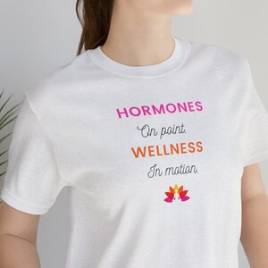 May include: White t-shirt with the text "HORMONES On point. WELLNESS In motion." and a stylized image of a flower with pink, orange, and yellow petals.