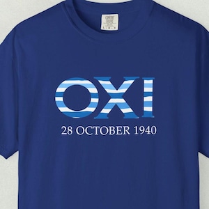 Könnte beinhalten: Ein königsblaues Kurzarm-T-Shirt mit dem Schriftzug OXI in großen, fetten Buchstaben mit einem horizontalen hellblau-weißen Streifenmuster. Unter dem Wort OXI ist der Text 28 OCTOBER 1940 in einer klaren, weißen serifenlosen Schrift gedruckt.