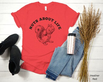 May include: Heather red t-shirt with the text "NUTS ABOUT LIFE" and a black illustration of a squirrel. The shirt is paired with blue jeans, black shoes, and a patterned travel mug. Dried wheat stalks are also in the image.