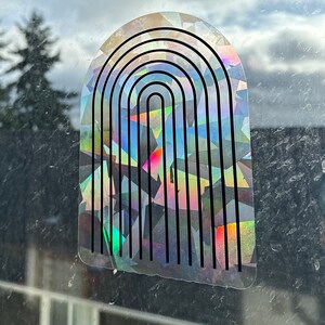 May include: A holographic rainbow arch sticker with a black outline. The sticker is made of iridescent material and has a textured surface.