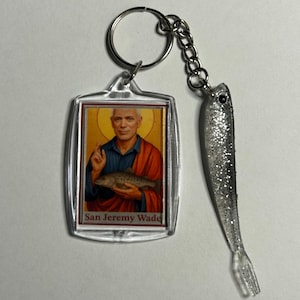 Peut inclure: Un porte-clés en acrylique transparent avec une image de San Jeremy Wade tenant un poisson, avec du texte en dessous. Il est orné d'un charm en forme de poisson argenté. Le porte-clés a un anneau et une chaîne argentés.