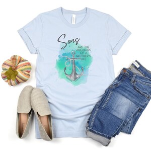May include: A pale blue t-shirt featuring the text "Sons are the anchors of a mother's life" and an anchor design. The image includes blue jeans, beige slip-on shoes, and a decorative pumpkin.