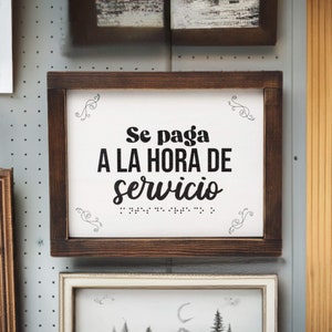 May include: A brown wooden framed sign with white background and black text that reads "Se paga a la hora de servicio" with braille dots below the text. The sign has decorative swirls in the corners.