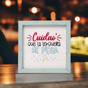 May include: A white wooden sign with a blue frame, featuring the text "Cuidado: que la loquera se pega" in red and black lettering. The sign is decorated with two champagne glasses clinking together.