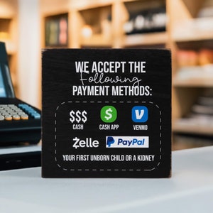 May include: Black chalkboard sign with white text that reads "WE ACCEPT THE Following PAYMENT METHODS: $$$ CASH, $ CASH APP, V VENMO, Zelle, PayPal, YOUR FIRST UNBORN CHILD OR A KIDNEY".