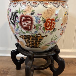 May include: A large, round, white ceramic fishbowl planter featuring a colourful hand-painted floral design with peonies, a woven basket motif, and a decorative border along the top rim. The bowl rests on a dark brown, carved wooden display stand.
