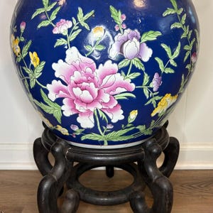May include: A large, round, cobalt blue ceramic fishbowl planter featuring hand-painted pink, purple, and yellow floral motifs with green leaves. The bowl has a white interior rim with small floral accents and rests on a dark brown wooden stand.