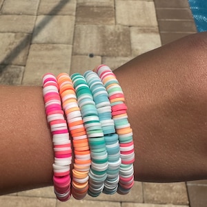 May include: A stack of colorful bracelets made of small, round beads. The bracelets are layered on a person's wrist and feature a variety of pastel colors, including pink, blue, green, and orange.