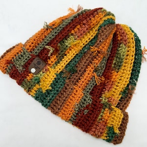 May include: A crocheted beanie featuring vertical stripes in shades of orange, brown, yellow, and dark green. The hat has a textured, frayed appearance with loose yarn ends throughout and a small brown leather tag labeled Dally Crochet Crafts.