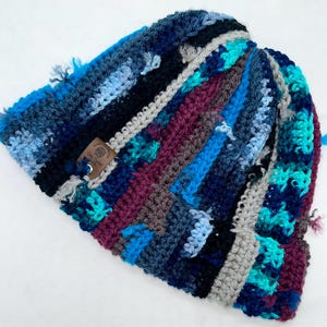 May include: A crocheted beanie featuring a multi-colored striped pattern in shades of navy blue, turquoise, light blue, grey, and deep burgundy. The hat includes a small, square brown leather tag with the text "Duly Crafted" attached to the side.