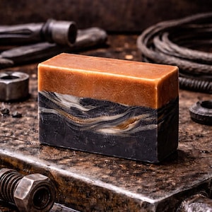 May include: A rectangular bar of soap with a layered design. The top layer is a warm brown, followed by a marbled white and gold layer, and a dark charcoal base. The soap is surrounded by metal tools.