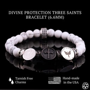 May include: White Howlite bead bracelet with three silver charms. The charms feature a cross, a Saint Michael design, and a Saint Benedict medal. The bracelet is 6.6mm wide and is tarnish free.