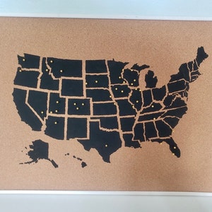 May include: A corkboard with a black outline of the United States map. Gold push pins are placed throughout the map, indicating different locations.