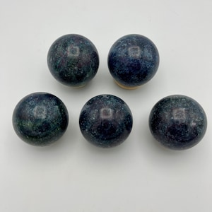 May include: Five dark blue-green decorative spheres with a speckled pattern. The spheres are arranged on a white surface. The spheres have a polished, smooth appearance and are likely made of stone or a similar material.