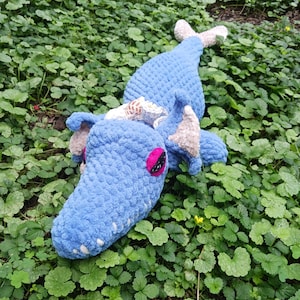 May include: A blue crocheted sea creature with a shell on its head. The creature has pink eyes and a white crocheted tail. It is lying on a bed of green leaves.