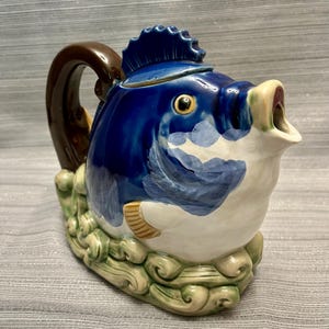 May include: A ceramic teapot shaped like a fish with a glossy dark blue body, a white underbelly, and a brown handle. The fish features a prominent dorsal fin lid, a wide open mouth spout, and is resting on a base sculpted to resemble green waves.