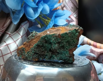 May include: A rough, green and brown malachite rock with a textured surface. The rock is sitting on a silver bowl.