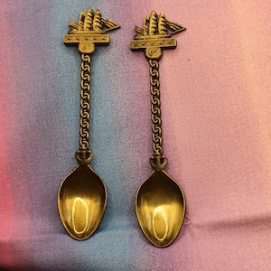 May include: Two antique gold-colored spoons with a ship design at the top and a chain-like handle. The spoons have an oval bowl shape. The background is a blend of pink, purple, and blue fabric.