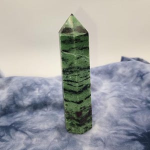 May include: A tall, green zoisite crystal obelisk with black and red inclusions. The polished stone has a pointed top and a rectangular base. The obelisk is standing upright against a light background.