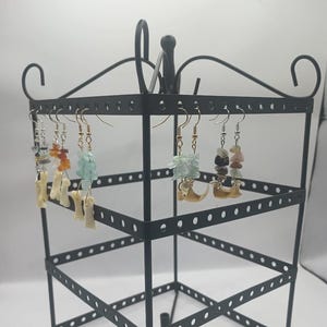 May include: A black metal earring display stand holding various handmade earrings. The earrings feature colorful gemstones, bone-shaped charms, and gold-colored hooks. The gemstones include blue, orange, and pink hues. The stand has multiple tiers for displaying jewelry.