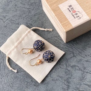 Puede incluir: Pendientes hechos a mano con diseños geométricos en azul marino y dorado. Los pendientes tienen ganchos dorados y una pequeña cuenta redonda dorada. Se exhiben en una bolsa blanca junto a una caja de madera con texto japonés.