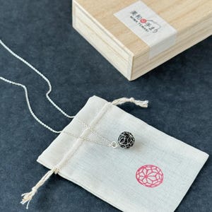 以下が含まれることがあります： 黒い透かし彫りの球体ペンダントが付いたシルバーネックレスが、小さな白い巾着袋の上に置かれています。背景には、日本語のテキストと赤い花柄模様が入った木箱があります。チェーンは繊細です。