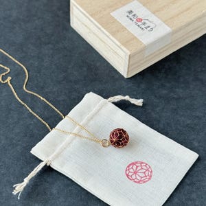 以下が含まれることがあります： 幾何学模様の赤い球体ペンダントが付いたゴールドカラーのネックレスが、白い巾着袋の上に置かれています。背景には日本語のテキストが入った木箱があります。ペンダントは球体です。