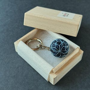 Könnte beinhalten: Ein handgefertigter Schlüsselanhänger mit einem blau-weißen, geometrisch gemusterten Ball. Der Schlüsselanhänger wird in einer kleinen, offenen Holzkiste mit hellem Innenraum auf dunklem, grauem Hintergrund präsentiert.