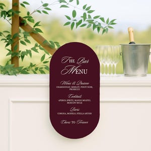 May include: A bar menu sign with a burgundy background lists wines, cocktails, and beers. The sign is next to a champagne bucket, glasses, and a cream-colored umbrella. Green leaves and a wooden pole are also visible.