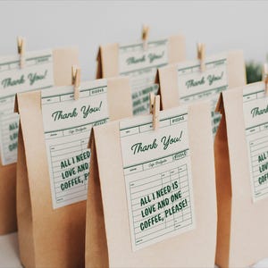May include: Several brown paper bags with tags that read "Thank You!" and "All I need is love and one coffee, please!" The tags are attached with small wooden clips. The bags are arranged on a white surface.