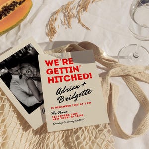 May include: A tan wedding invitation with red text that reads "We're Gettin' Hitched!" with the names Adrian and Bridgette. The date and time of the wedding is December 15th at 3 PM. The venue is 123 Lovers Lane, New York, NY 12345. The invitation says "Drinking & dancing to follow!"