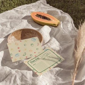 May include: Two recipe cards with a fruit pattern on a white tablecloth. The top card has a pattern of lemons, strawberries, watermelon slices, and tomatoes. The bottom card has the text "Recipe Card" and a blank space for writing ingredients and instructions. A papaya half and a brown bowl are also visible.